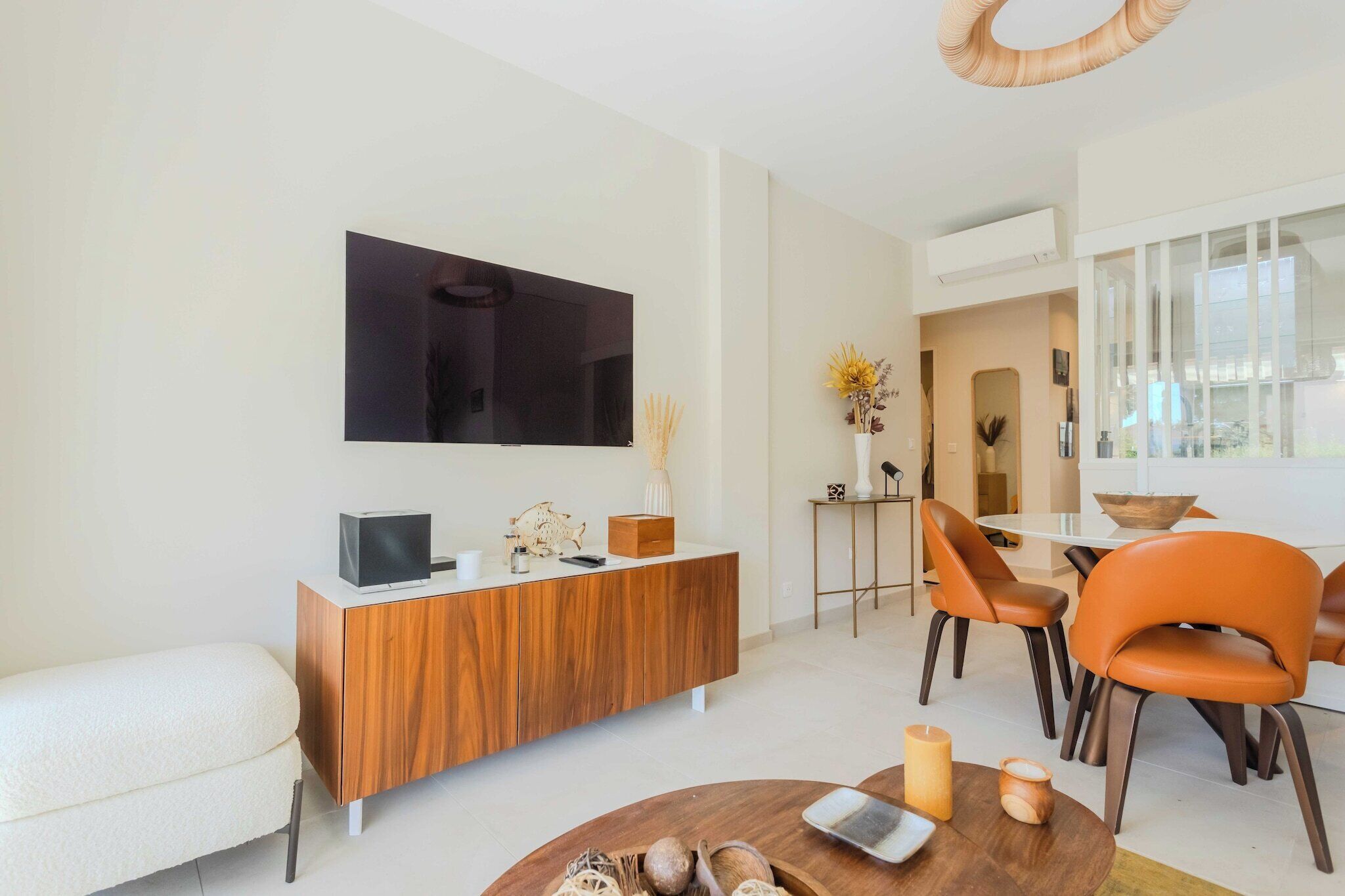 A modern living room with a flat-screen TV, wooden console, orange chairs, and a dining area with a glass table.
