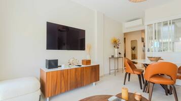 A modern living room with a flat-screen TV, wooden console, orange chairs, and a dining area with a glass table.