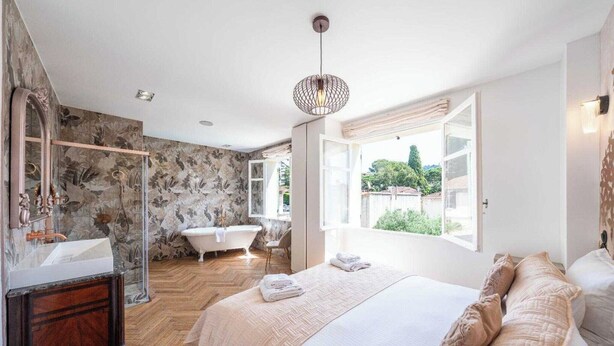 Ein Schlafzimmer mit einem großen Bett, ein Badezimmer mit einer Badewanne und Blick auf Grünflächen.