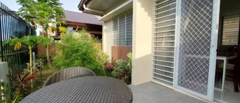 A patio with a round wicker table and chairs, a sliding glass door with a metal security screen, and a garden with various plants and a fence.