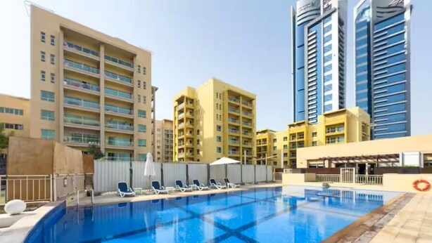 Una piscina con sillas de descanso y sombrillas, rodeada de edificios de varios pisos.
