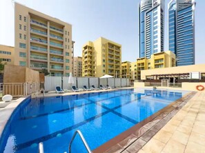 A swimming pool with lounge chairs and umbrellas, surrounded by multi-story buildings. - Vogue | Al Alka Greens near Mall of the Emirates (Dubai)