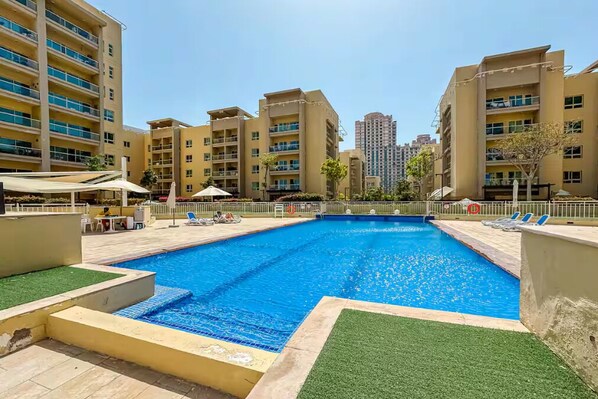 A swimming pool surrounded by multi-story buildings with balconies, a covered seating area, and a clear sky. - Vogue | Al Alka Greens near Mall of the Emirates (Dubai)