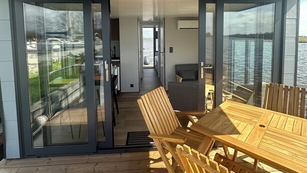 A modern outdoor dining area with a wooden table and chairs, a glass-enclosed space, and a view of the water. - Super nice houseboat with fantastic views! (Molkwerum)