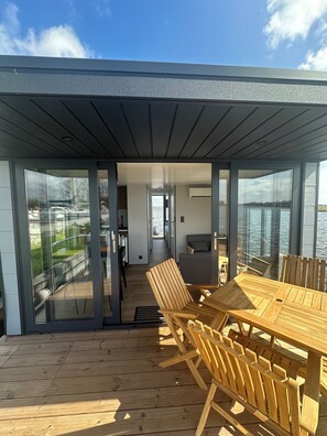 A modern outdoor dining area with a wooden table and chairs, a glass-enclosed space, and a view of the water.