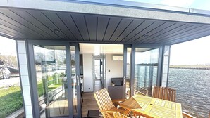 A modern outdoor deck with a glass enclosure, wooden furniture, and a view of the water. - Super nice houseboat with fantastic views! (Molkwerum)