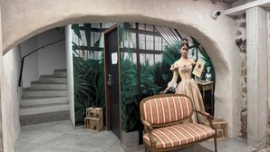 A room with a staircase, a decorative chair, and a mannequin dressed in a vintage gown.