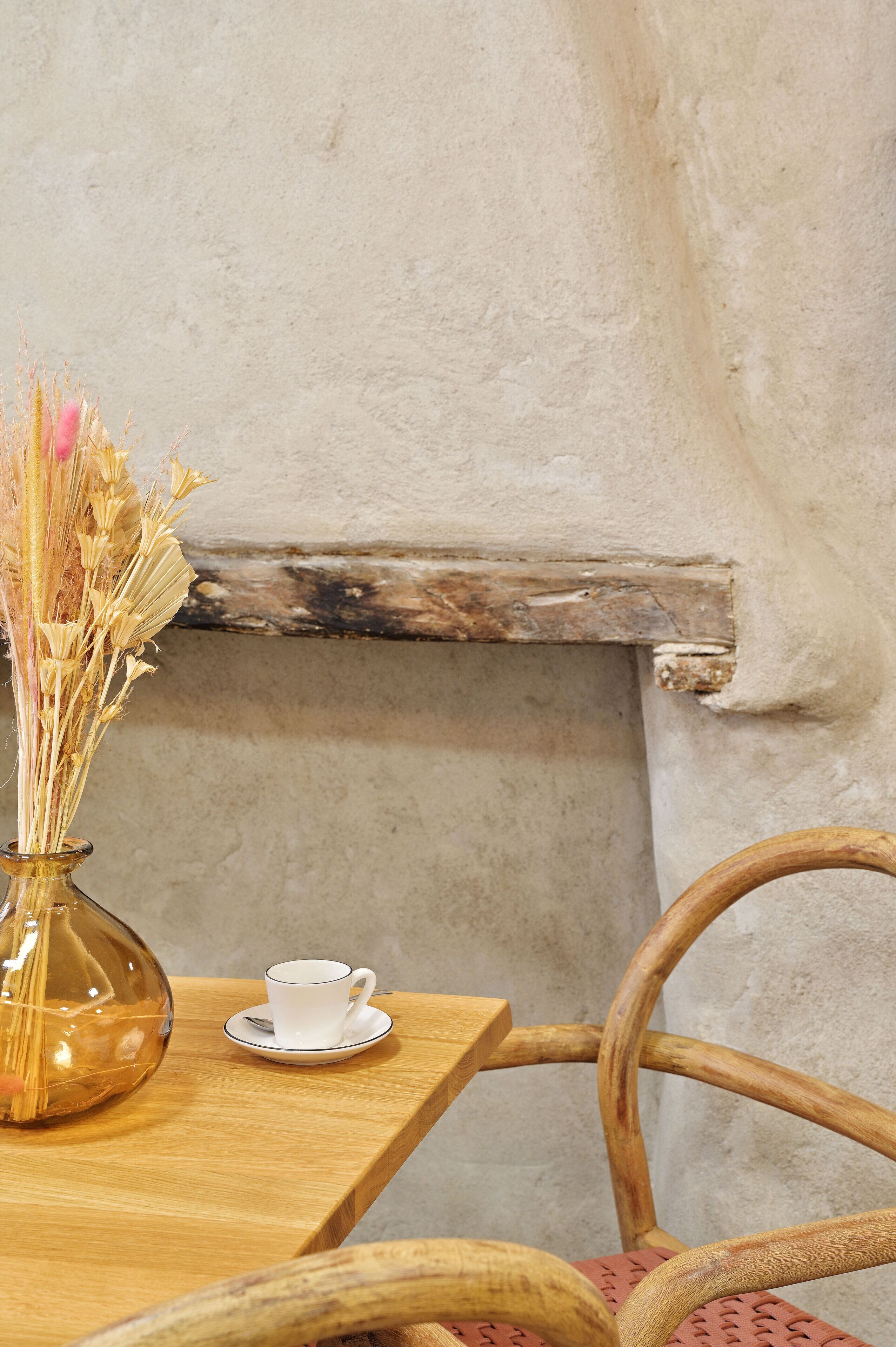 A wooden table with a clear glass vase containing dried flowers, a white cup, and a wooden chair.