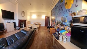 A living room with a black leather sofa, a flat-screen TV, a fireplace, a dining area with a table and chairs, and a kitchen area with a microwave and various containers. - Crittlewood Guest House (Gainsborough)