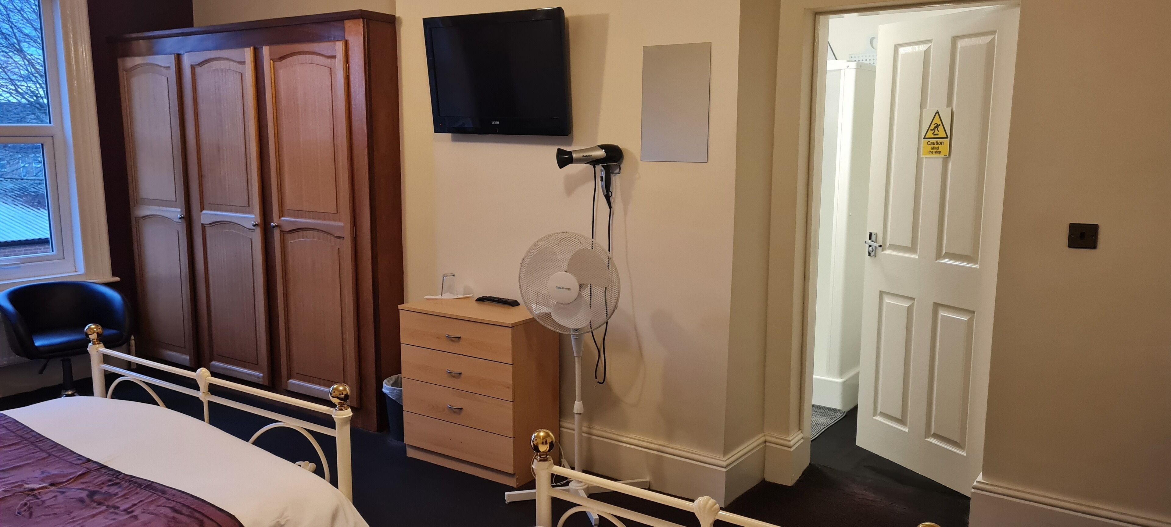A bedroom with a bed, wooden wardrobe, a television, a fan, and a door with a caution sign.