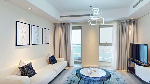 A modern living room with a white sofa, a round coffee table, and a flat-screen TV. There are three framed artworks on the wall and a large window with curtains. - Luxurious 1 Bedroom-High floor-Sea view and Balcony (Dubai)