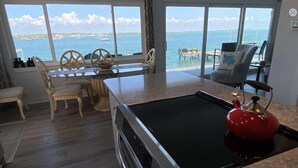 A kitchen with a large window overlooking the sea, a red kettle, and a dining area with wooden chairs.