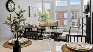 Un comedor con una mesa preparada para dos personas, un reloj en la pared y un sofá al fondo.