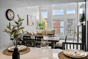 A dining area with a table set for two, a clock on the wall, and a sofa in the background. - 3BR | Garage | DT Views | Central | Lux Townhome (Calgary)