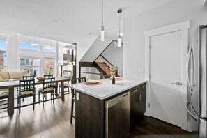 A modern kitchen with a central island, stainless steel appliances, and a dining area with wooden furniture. - 3BR | Garage | DT Views | Central | Lux Townhome (Calgary)