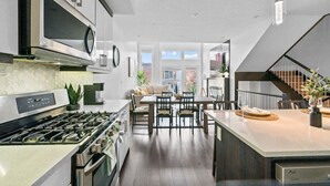 Una cocina moderna con electrodomésticos de acero inoxidable, un comedor con mesa y sillas de madera, y una escalera que sube a un nivel superior.