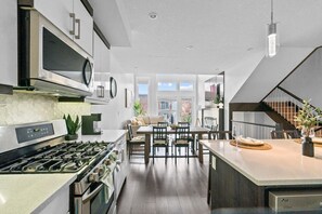 A modern kitchen with stainless steel appliances, a dining area with a wooden table and chairs, and a staircase leading to an upper level. - 3BR | Garage | DT Views | Central | Lux Townhome (Calgary)