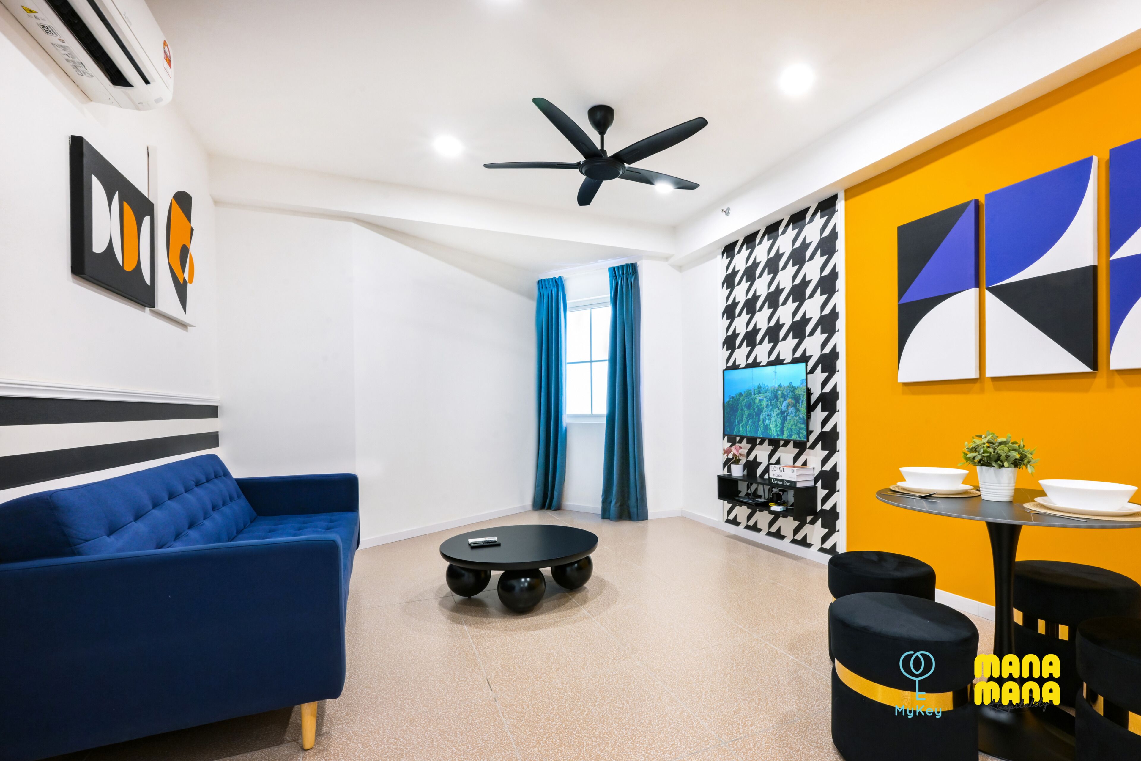 Een moderne woonkamer met een blauwe bank, een ronde tafel met krukken, een plafondventilator en kunst aan de muur.