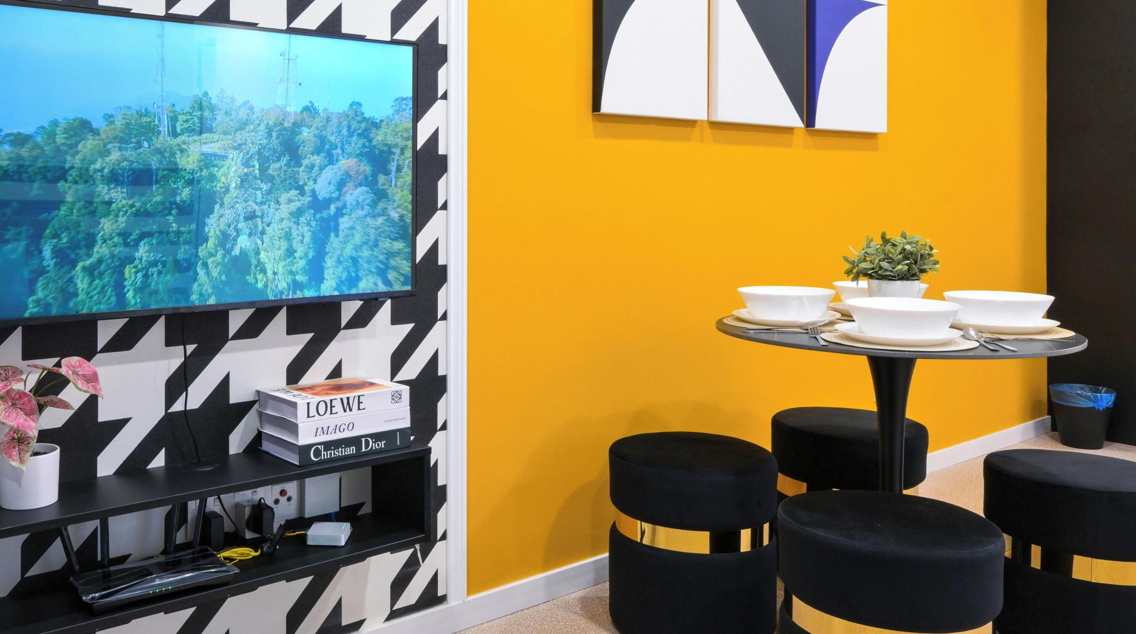 A modern hotel room with a large TV mounted on a black and white chevron-patterned wall, a small dining table with white bowls and a potted plant, and black ottomans with gold stripes.