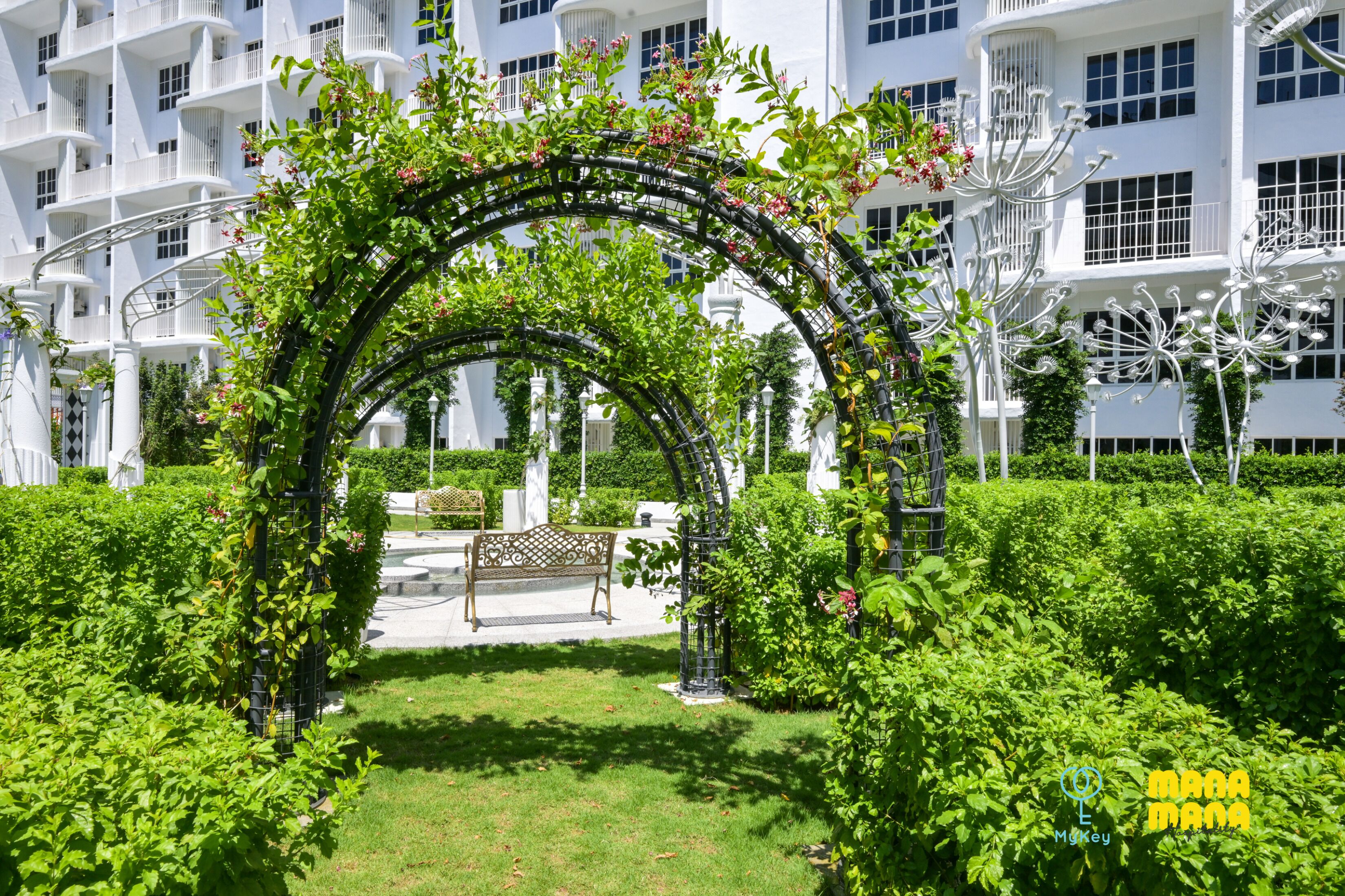 Un jardin agrémenté d’une tonnelle en arc de cercle, d’un banc et, au fond, d’un bâtiment blanc doté de balcons.