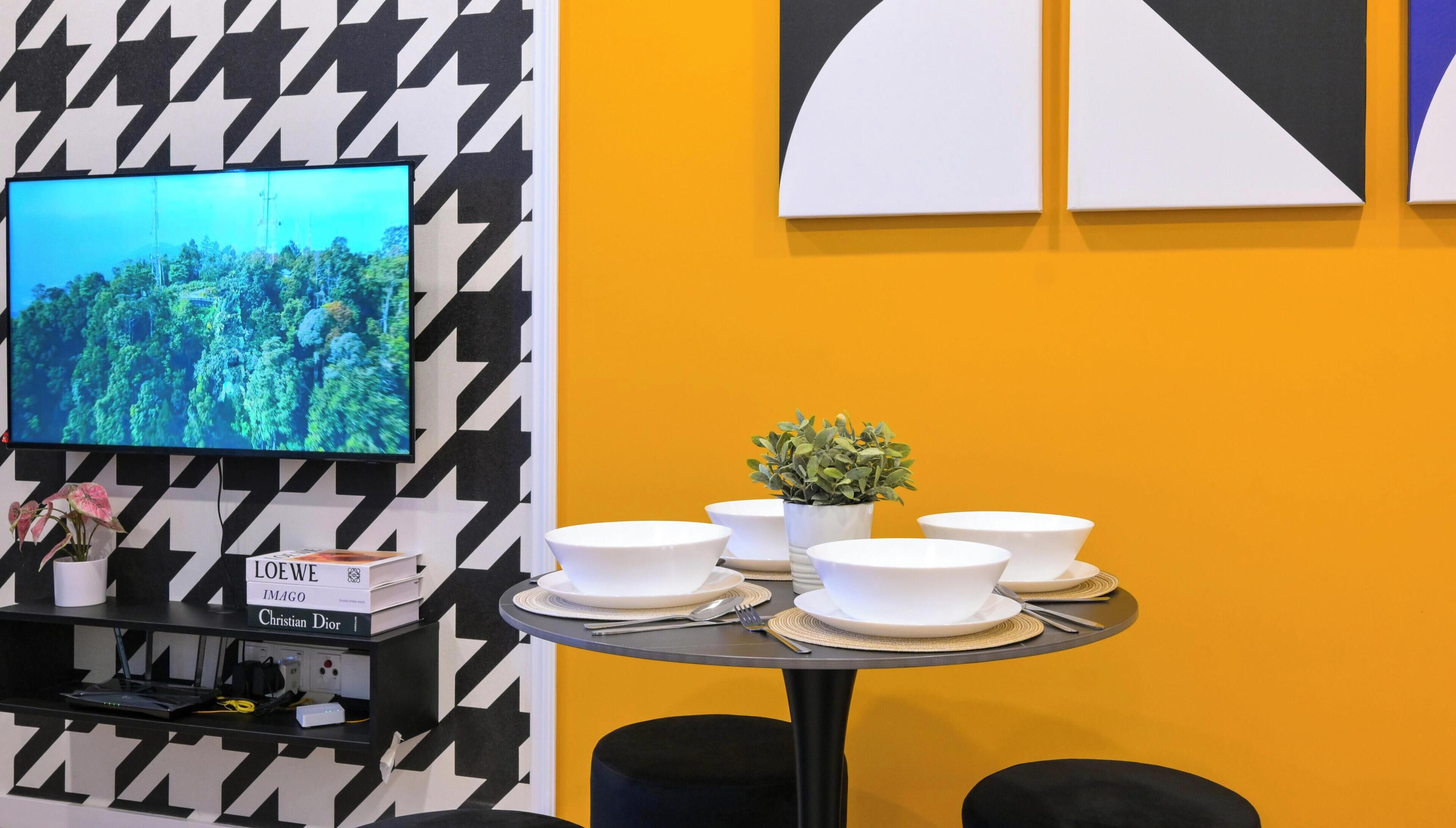 Un interior moderno con un televisor que muestra un paisaje boscoso, una mesita con cuencos blancos y una planta en maceta, y una estantería con libros y una placa con nombre.
