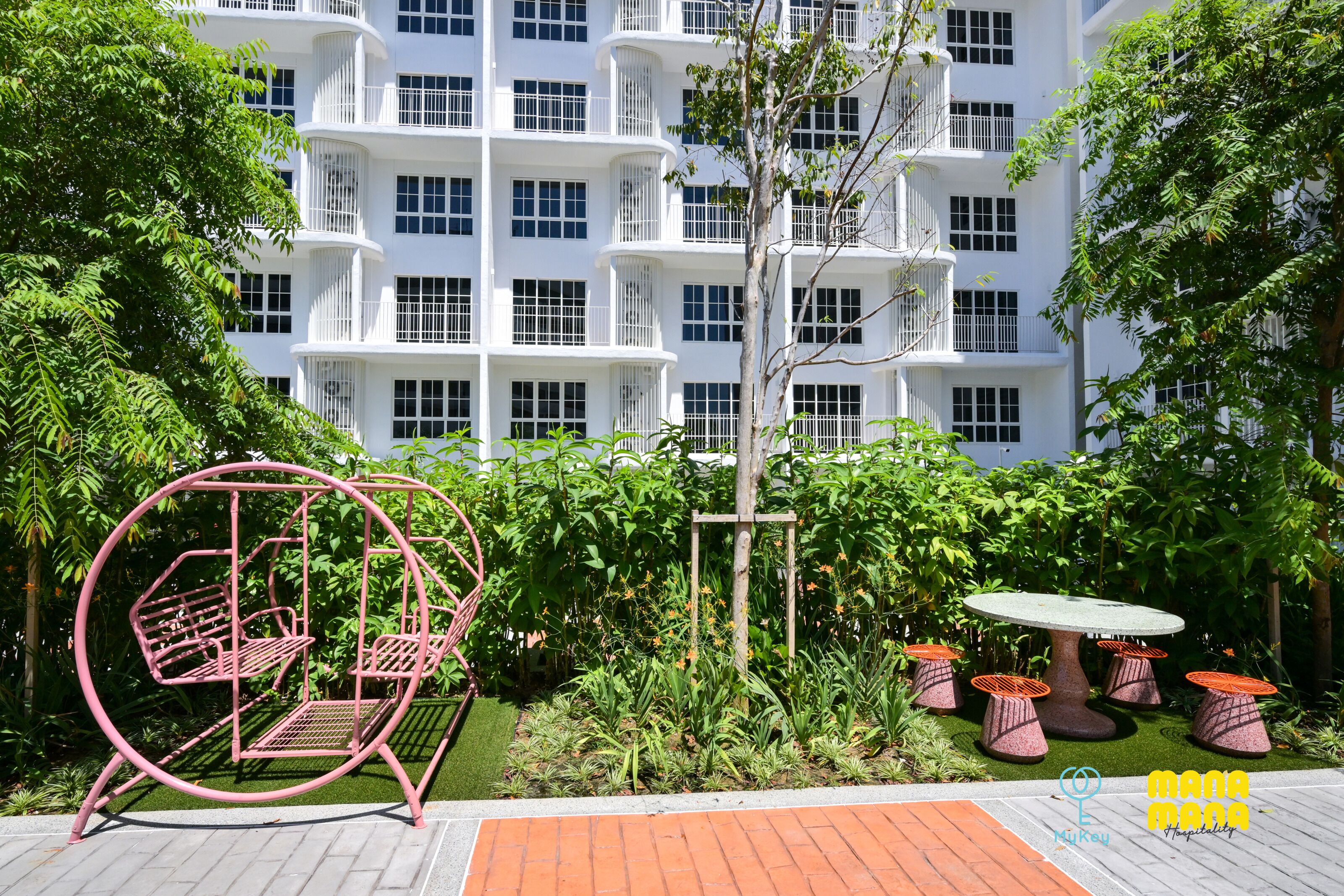 A garden area with a swing set, a small table with stools, and a building with balconies in the background.