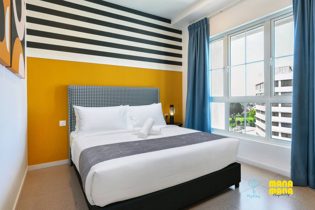 Una cama bien hecha con un cabecero a rayas, un ventanal con cortinas y vistas a edificios.