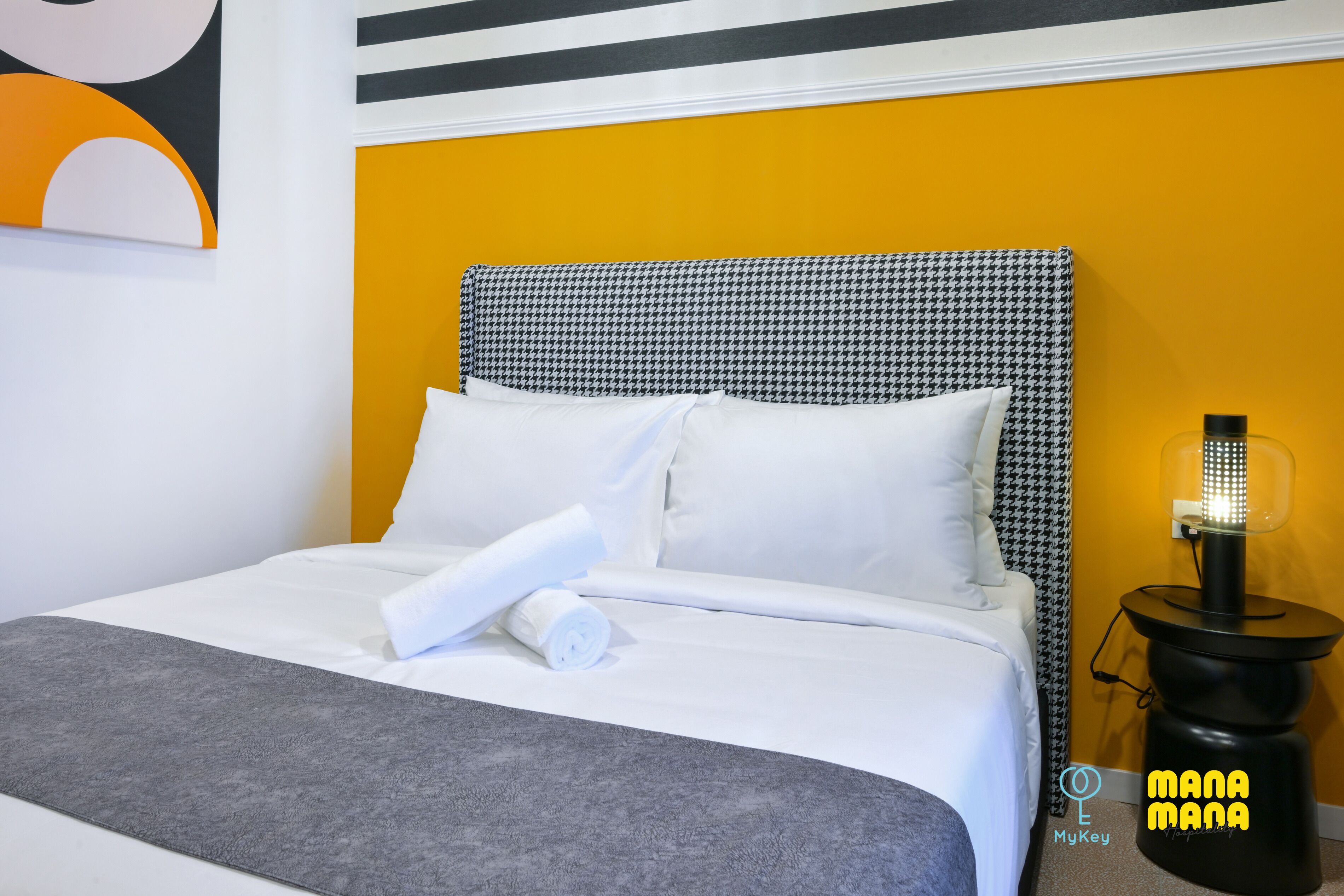 A neatly made bed with white linens, a grey throw blanket, and a black and white houndstooth headboard. A black nightstand with a lamp is visible to the right.