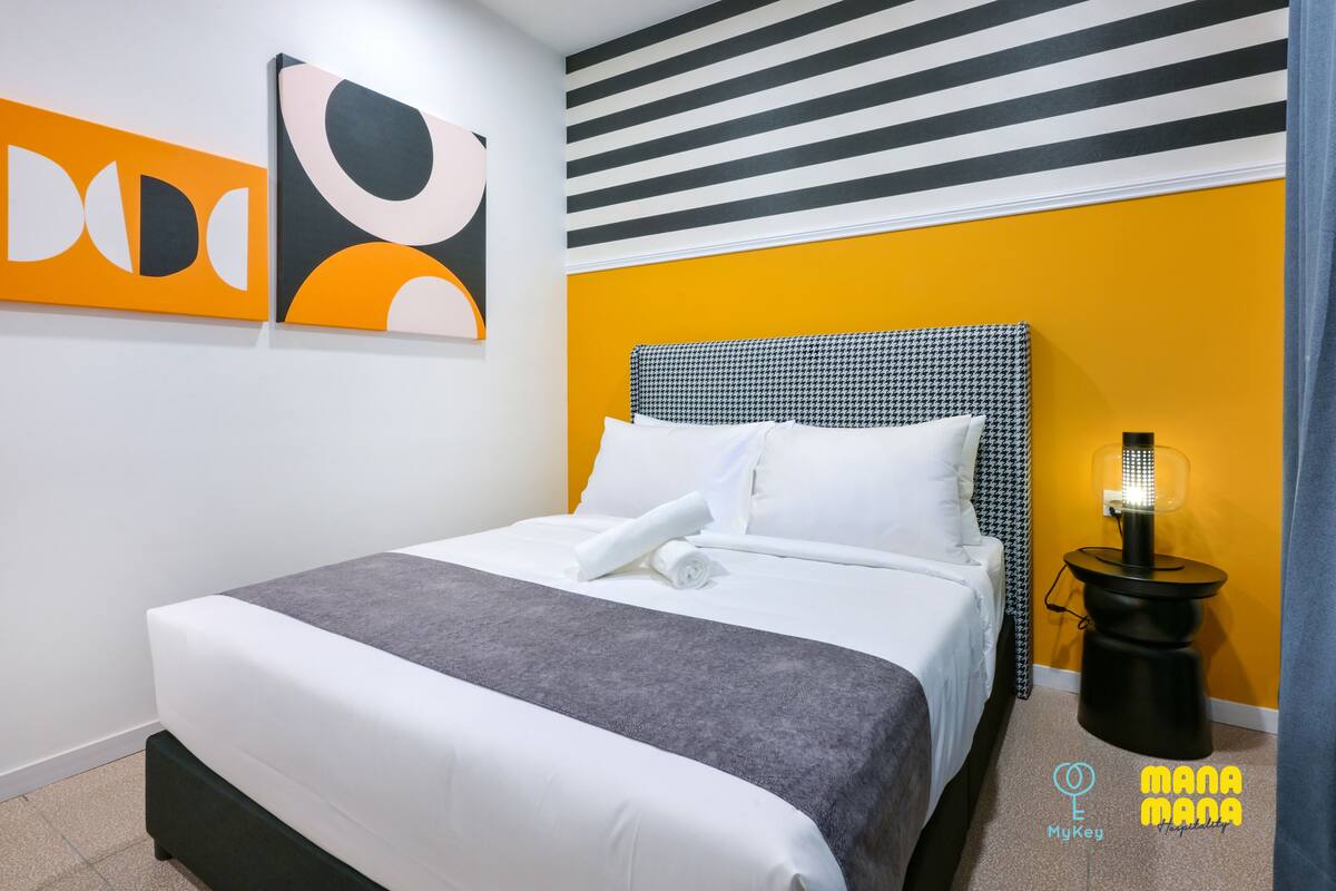 Una cama bien hecha con sábanas blancas y un plaid gris, un cabecero a cuadros negros y blancos estilo pied-de-poule y una mesita de noche negra con una lámpara.
