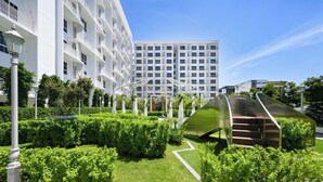 Een modern hotel met een witte gevel, meerdere balkons en een verzorgde tuin met groene hagen en een metalen trap.