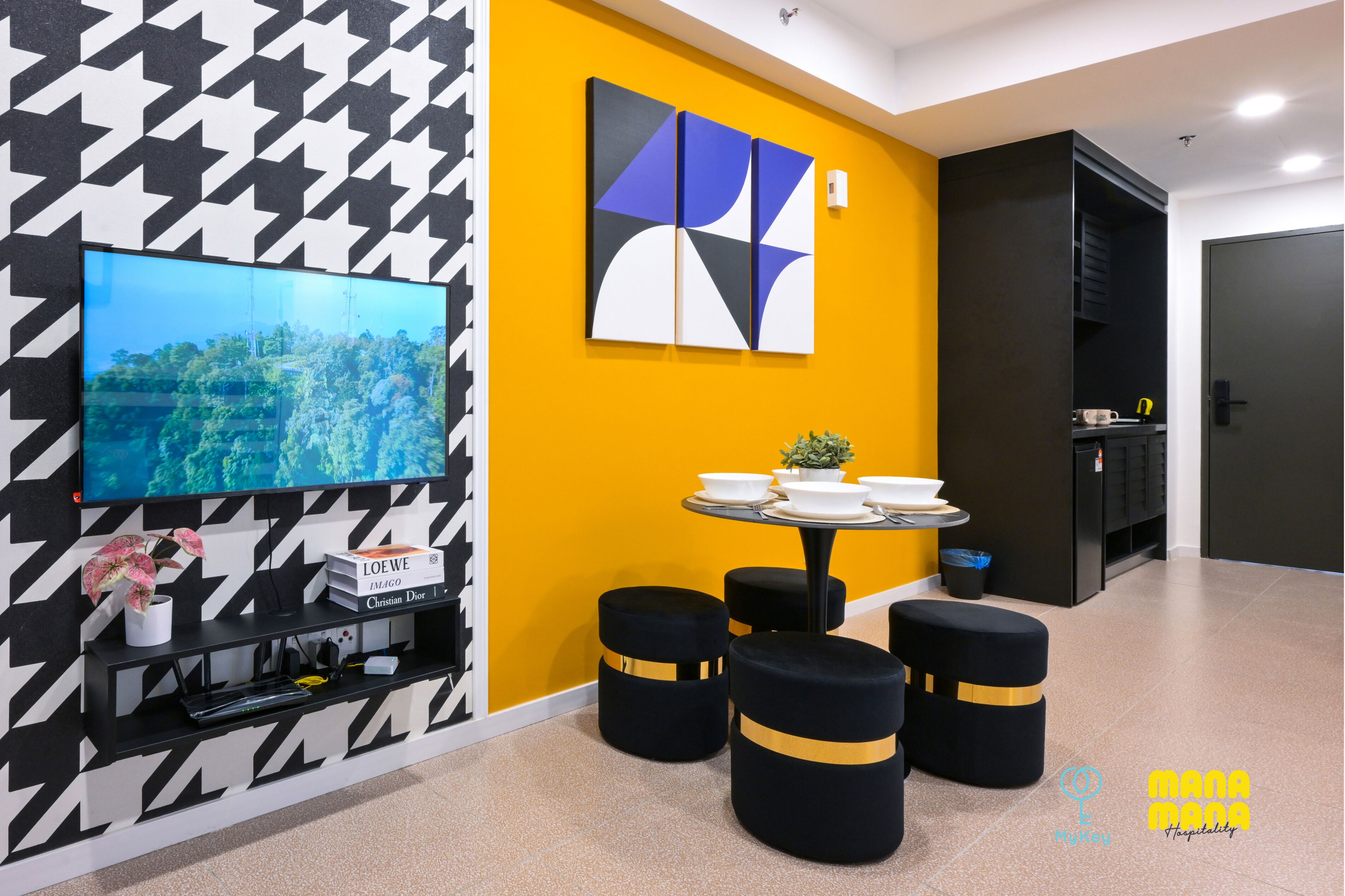A modern hotel room with a large flat-screen TV mounted on a black and white patterned wall, a round dining table with stools, and a yellow accent wall.