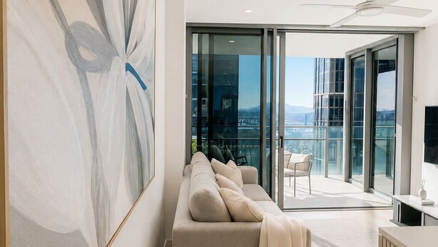 Una sala de estar moderna con un sofá, un cuadro abstracto de gran tamaño y vistas a un paisaje urbano a través de amplios ventanales.