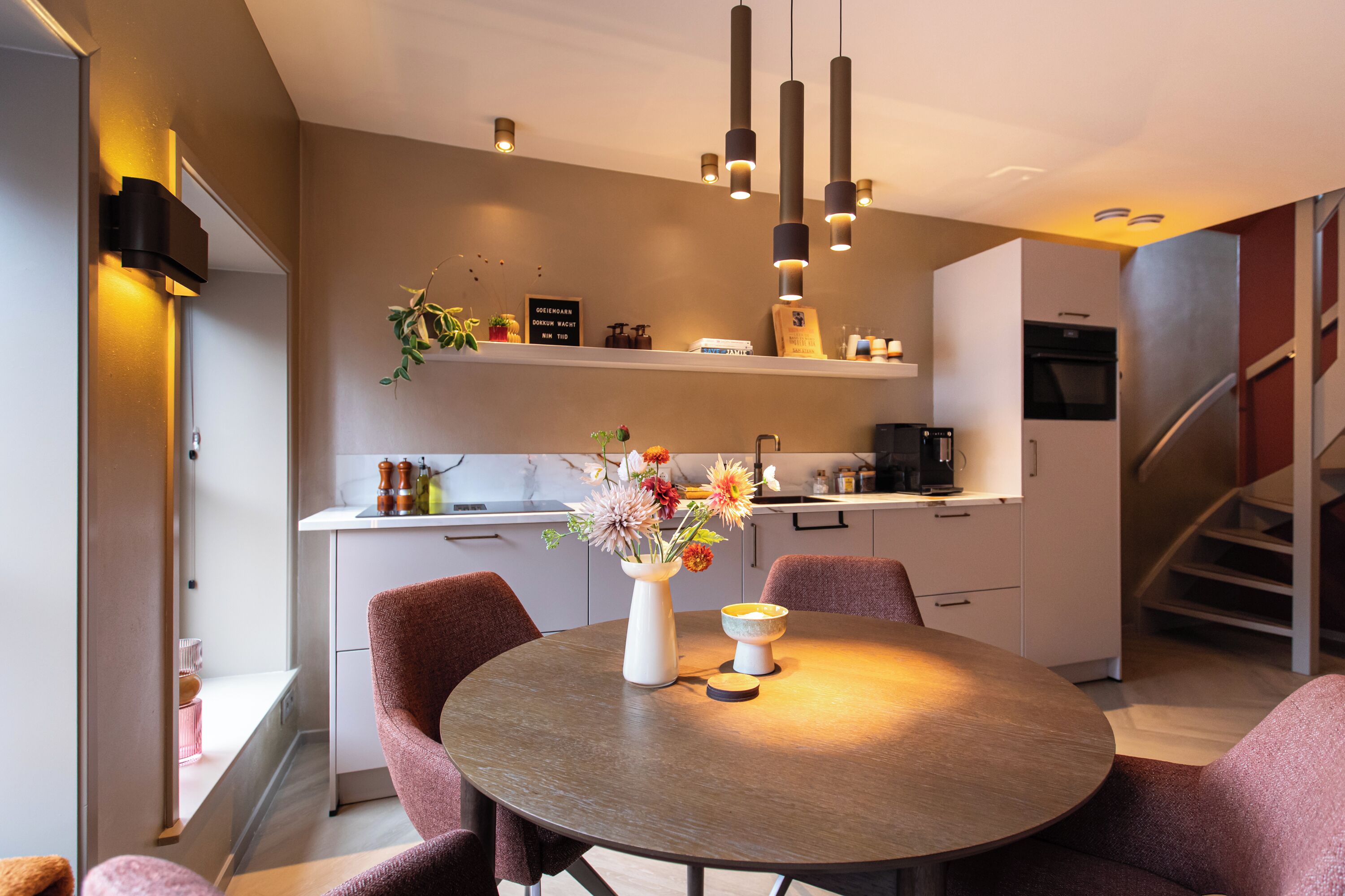A modern kitchen with a dining area, featuring a round table, chairs, pendant lights, and a staircase.