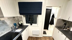 Una cocina estrecha con suelo de madera, armarios blancos, encimera negra y una ventana con persiana oscura.
