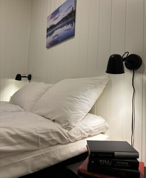 Una cama bien tendida con ropa blanca, una lámpara de pared y un cuadro enmarcado encima.