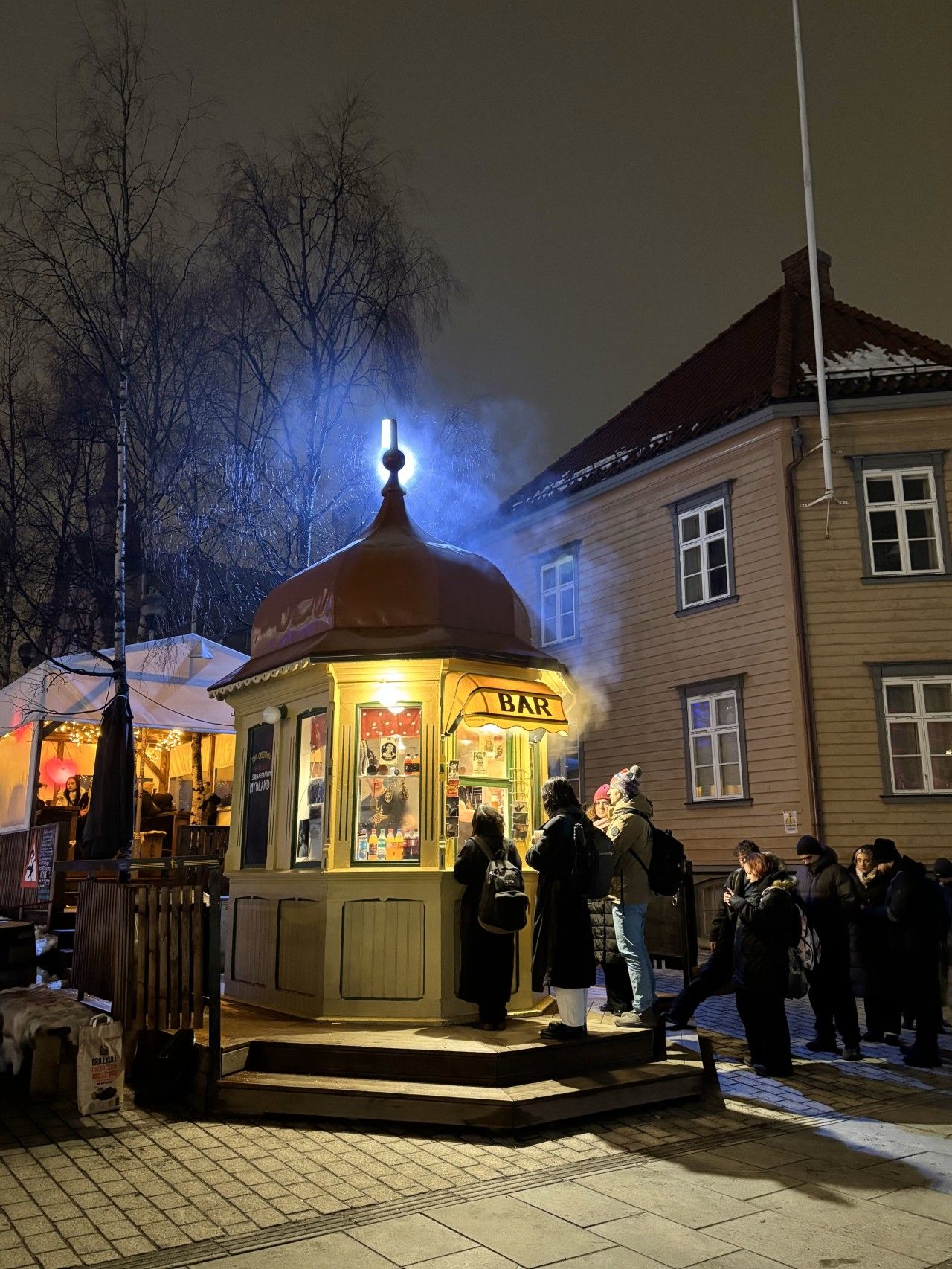 Pieni, valaistu kioski, jossa on kyltti, jossa lukee »BAR», ja sen ymp ärillä on ihmisiä sekä taustalla näkyy rakennus.