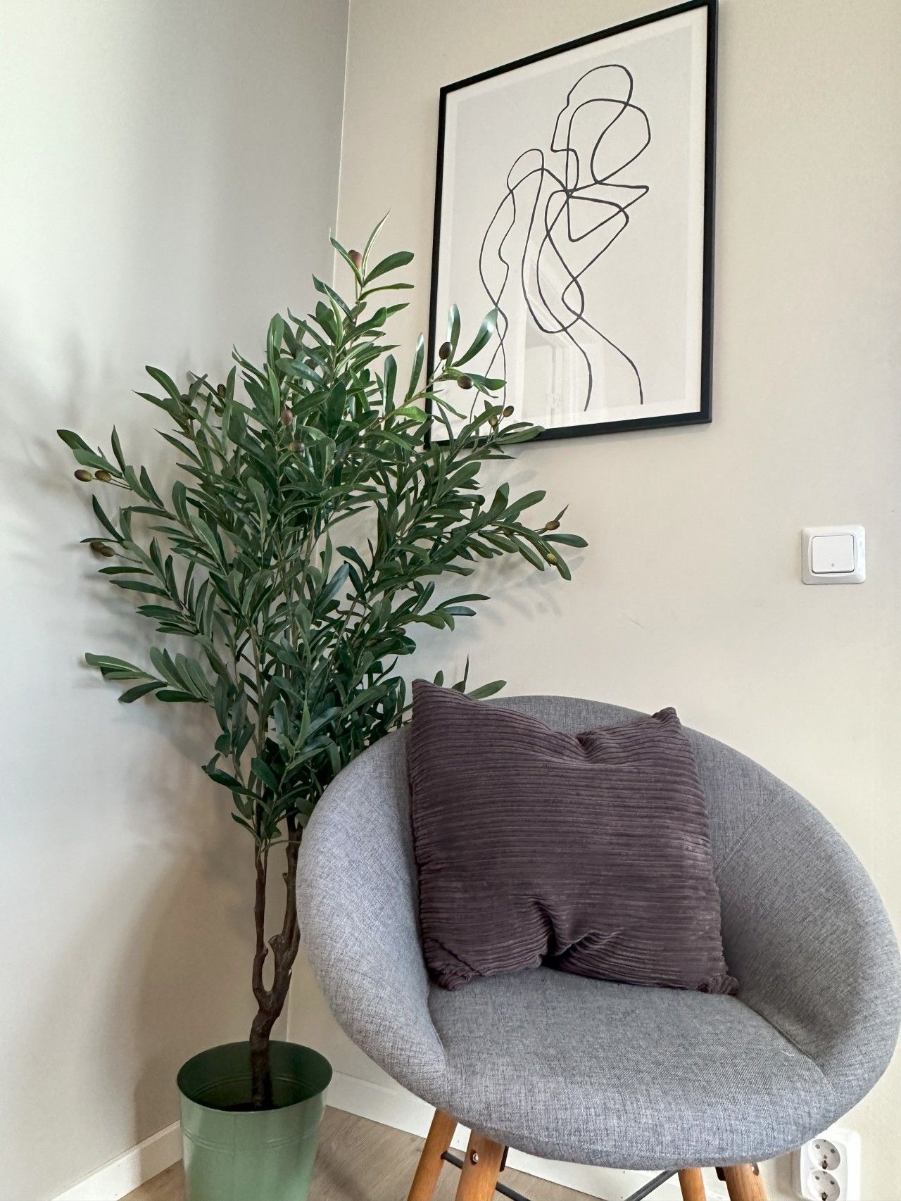 Une pièce avec un fauteuil gris, une plante verte dans un pot et une œuvre d’art abstraite encadrée accrochée au mur.