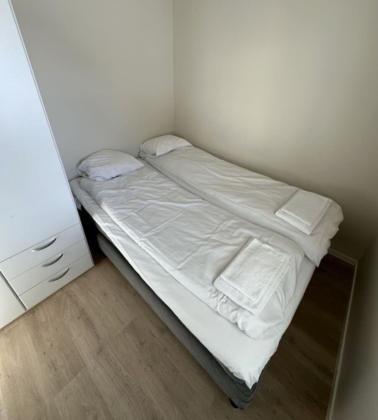 Piccola camera con un letto singolo, copripiumino, cuscino e coperta bianchi. Si vede un armadio bianco con un cassetto e un'anta per appendere. Il pavimento è in legno.