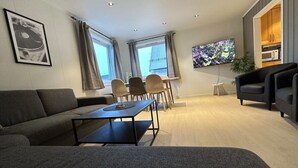 A modern living room with a sofa, a coffee table, a dining area with chairs, a TV mounted on the wall, and a kitchen area visible through an open door. - Sentrumsnær leilighet med 5 soverom - Montis (Tromsø)