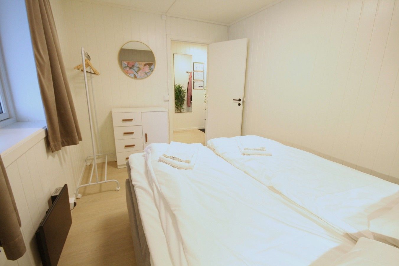 Una habitación con dos camas, un tocador blanco, un espejo redondo y una ventana con cortinas.