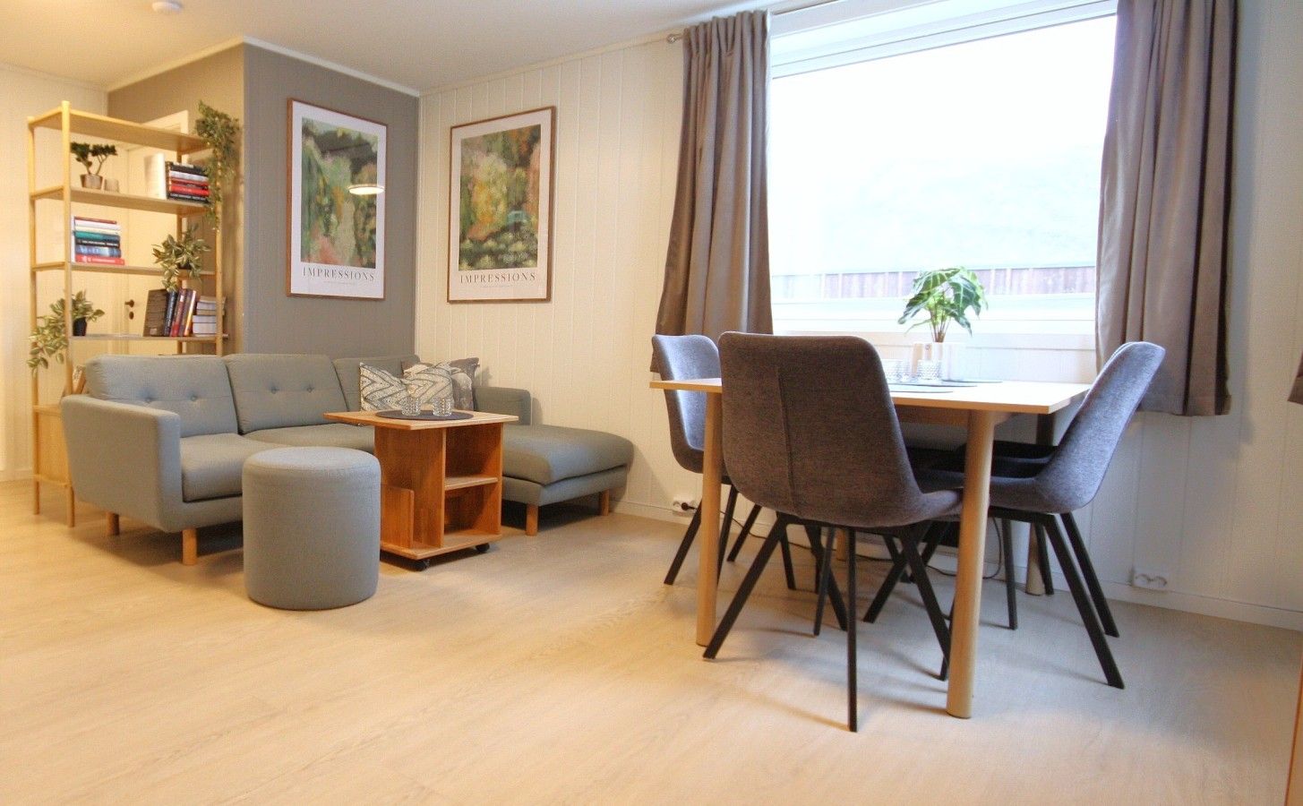 Een moderne woonkamer met een bank, voetenbankje, eettafel en stoelen. Er hangen ingelijste kunstwerken aan de muur en een boekenkast met boeken en planten.