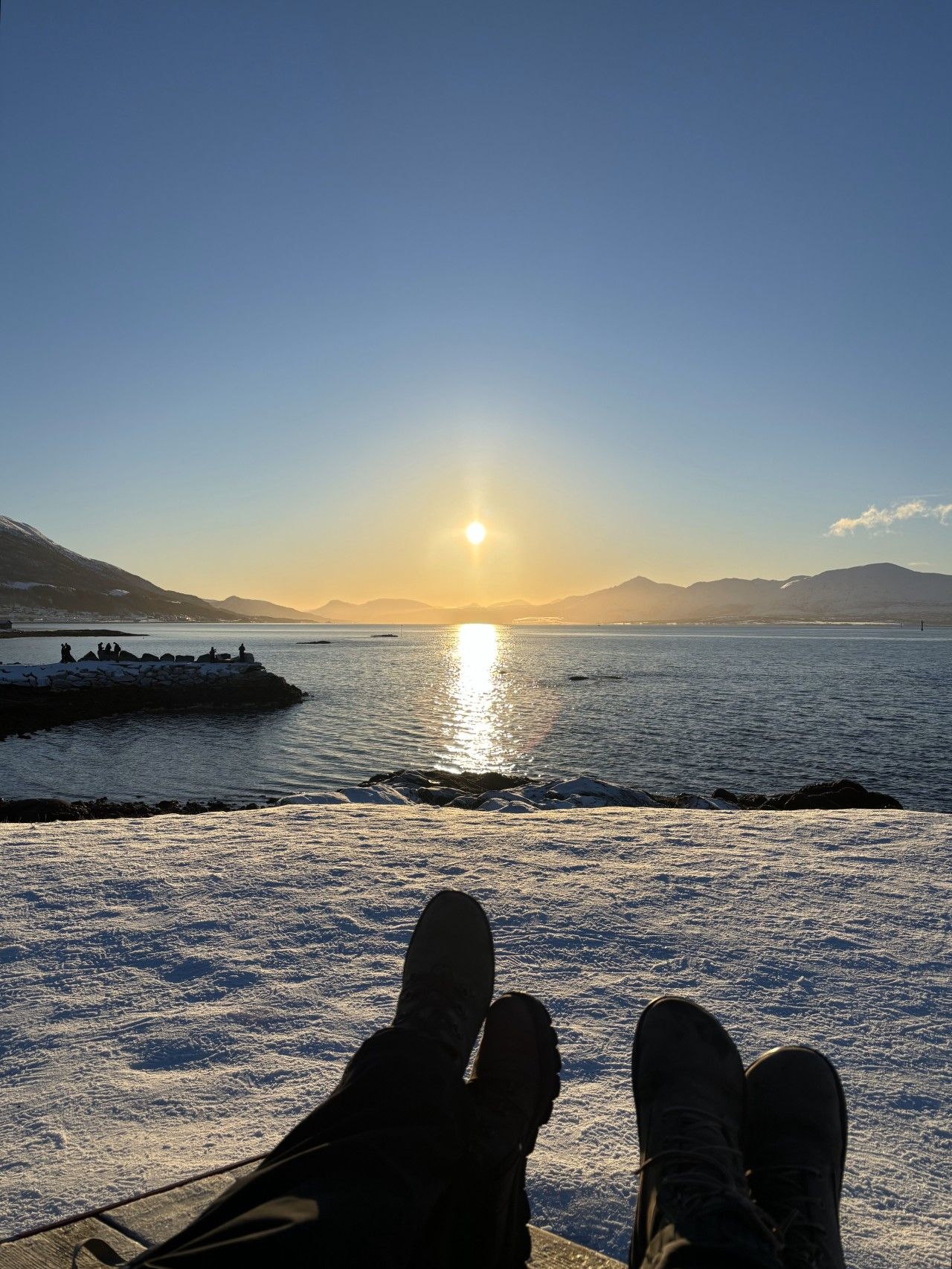 A person seated with their feet on a railing overlooking a body of water during sunset.