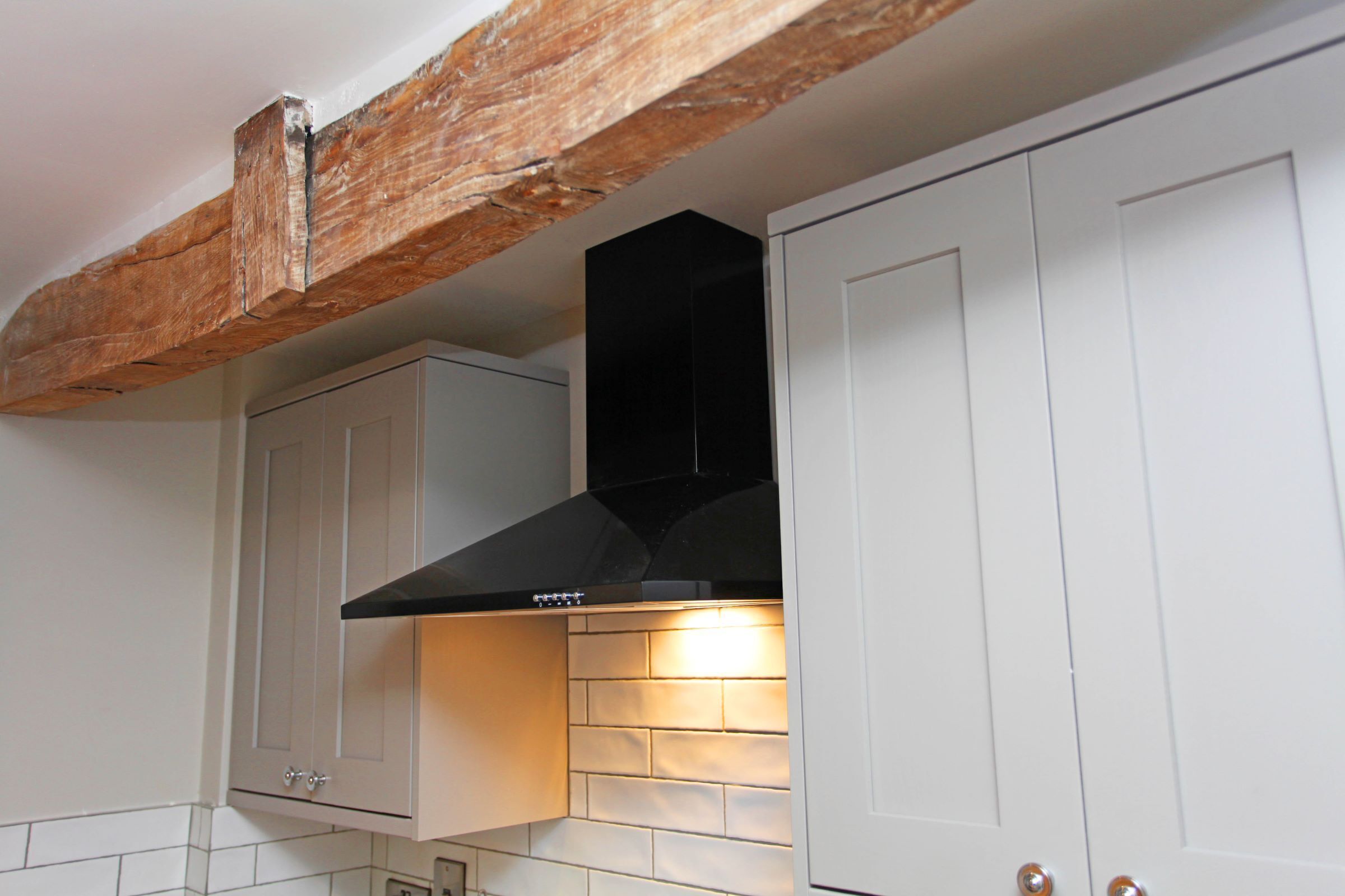 A kitchen with white cabinets, a black range hood, exposed wooden beams, and a tiled backsplash.