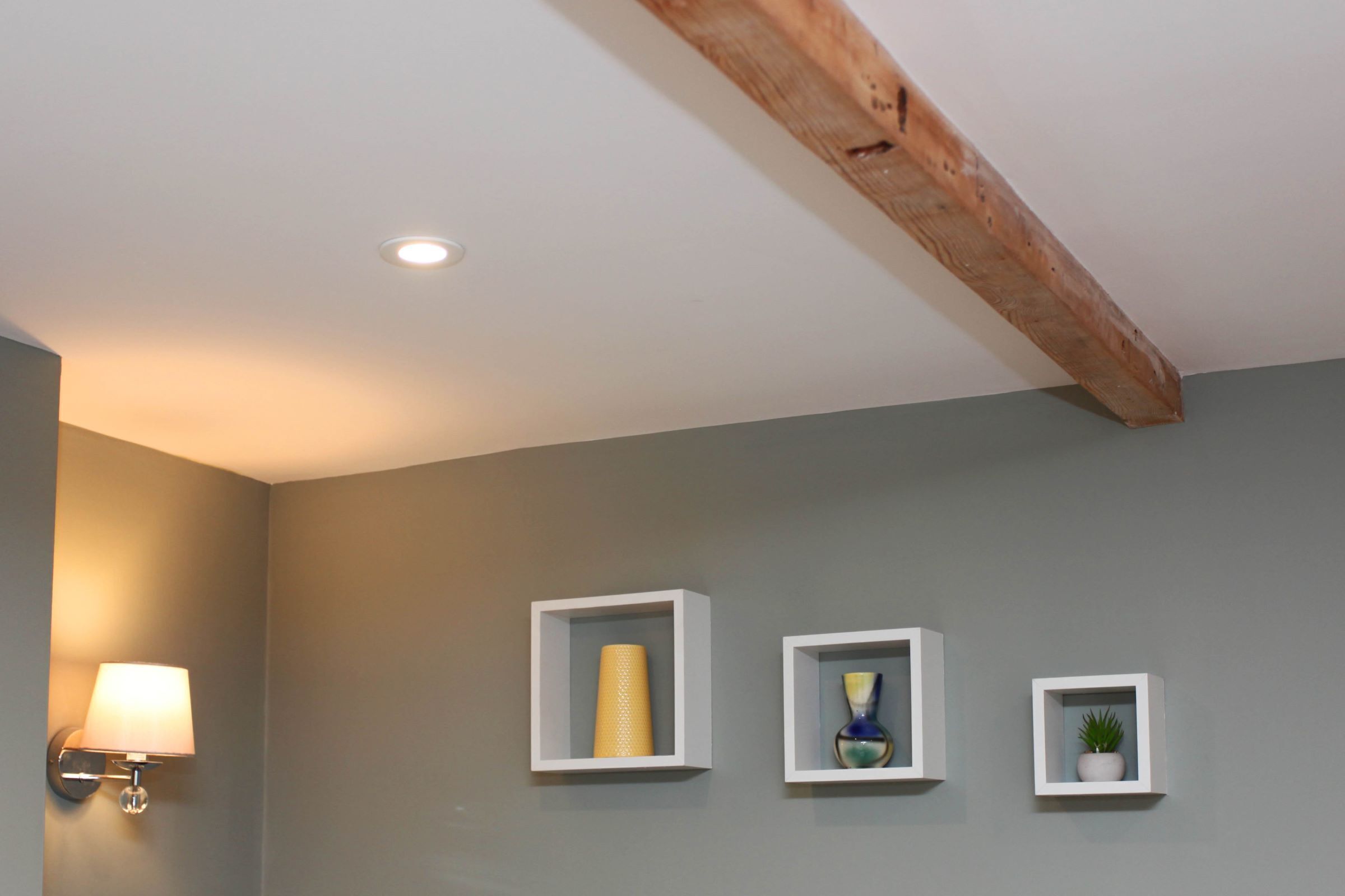 Une pièce avec un mur gris, un plafond avec une poutre en bois et trois étagères à cadre blanc ornées d’objets décoratifs.
