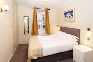 A bedroom with a bed, a nightstand, a dresser, a window with curtains, a mirror, and a painting on the wall. - St John's Hill Apartments (Shrewsbury)
