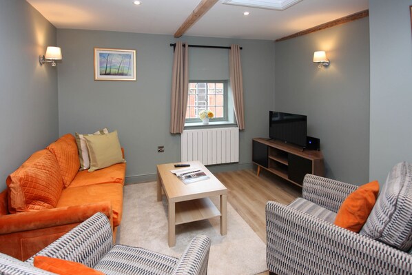 A living room with a sofa, armchairs, a coffee table, a TV, and a window with curtains. - St John's Hill Apartments (Shrewsbury)