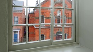 A window with a grid pattern, through which a red brick building with arched windows and a central circular window is visible.