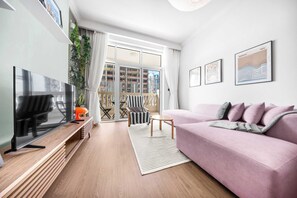 A modern living room with a flat-screen TV, a pink sofa, and a balcony with a view of buildings. - StayShort - Stylish 1-Bedroom Retreat in JVC that Sleeps 2 (JVC)