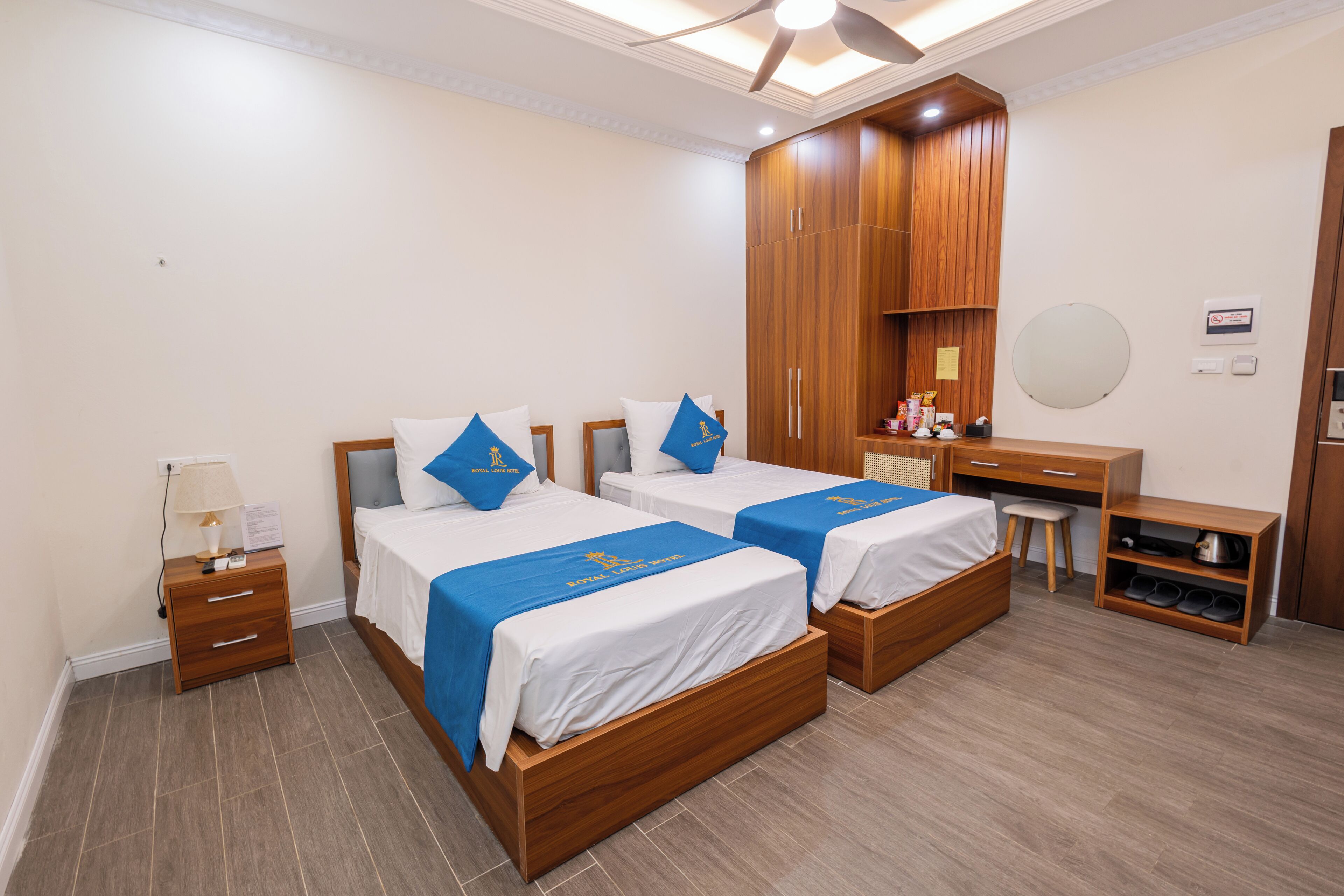 A hotel room with two wooden beds, a desk with a chair, a wardrobe, and a small shelf with shoes.