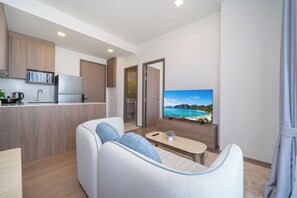A modern living room with a sofa, a coffee table, a TV displaying a scenic view, and a kitchen area with wooden cabinets and appliances. - Canvas Condo New 2BR Near Beach 111-80 (Choeng Thale)